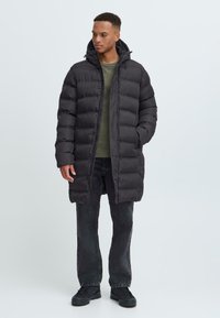 Abrigo de plumas negro con capucha, diseño acolchado y cierre a presión, llevado sobre un suéter verde y jeans oscuros, en un fondo claro.