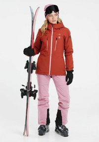 Rote wasserdichte Jacke mit pinken Akzenten, pinke Skihosen, schwarze Handschuhe und Skischuhe, hält pinke und schwarze Skier mit Bindungen.