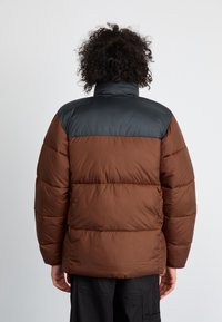 Chaqueta acolchada marrón y negra con cuello alto, que presenta secciones acolchadas y una textura suave en los hombros.