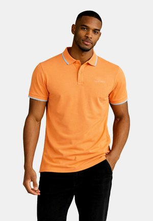 Mand iført en orange polotrøje med hvide kanter og tekst "California", stående med den ene hånd i lommen mod en ensfarvet baggrund.