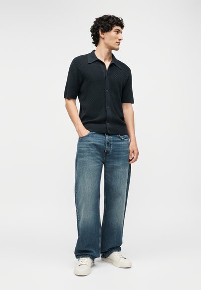 Νέος άνδρας με κατσαρά μαλλιά, φοράει μαύρο πουκάμισο με κοντά μανίκια και κουμπιά, μπλε φαρδιά τζιν και λευκά αθλητικά παπούτσια, στεκόμενος με τα χέρια στις τσέπες.
