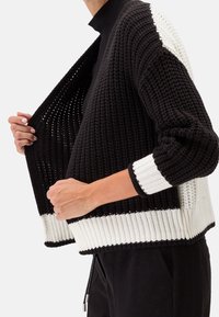 Svart och vit stickad cardigan med öppen fram, som har texturerade mönster och ribbade ärmslut. Designen kombinerar tät och mesh-sektioner.