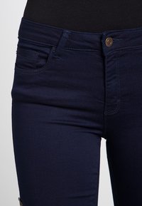 Jean en denim bleu foncé avec une coupe ajustée, présentant une fermeture à bouton à l'avant, des poches standard et une texture lisse.
