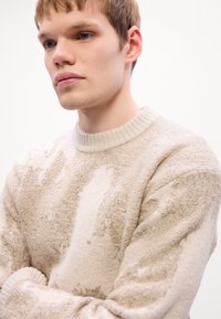 Young man with light skin and short hair wearing a beige textured sweater, looking forward with arms crossed against a white background.
