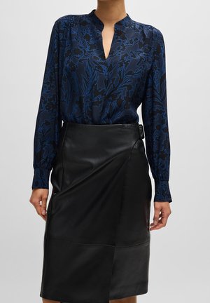 Kobieta ubrana w granatową bluzkę z długim rękawem i ciemnoniebieskim kwiatowym wzorem, włożoną do wysokiej taliowanej czarnej skóry spódnicy z boczną klamrą.