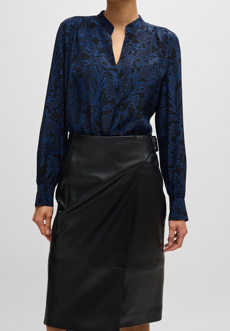 Femme portant un chemisier bleu marine à manches longues avec des motifs floraux bleu foncé, rentré dans une jupe en cuir noire taille haute avec une boucle sur le côté.