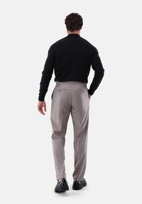 Mann, der von hinten steht, trägt ein schwarzes Langarmshirt, beige maßgeschneiderte Hose und schwarze Anzugschuhe vor einem weißen Hintergrund.