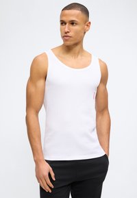 Μυώδης άνδρας που φοράει λευκό αμάνικο τοπ και μαύρο παντελόνι, στέκεται με το ένα χέρι στην τσέπη και κοιτάζει προς τα πλάγια.