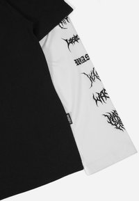 Zwart-witte lange mouw T-shirt met een zwart grafisch ontwerp op de witte mouw en een klein label met de tekst "WASTED."