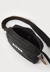 Čierna nylonová crossbody taška so zipsovým hlavným priehradkom, s hladkým povrchom a bielym brandingom. Nastaviteľný popruh s kontrastnými akcentmi.