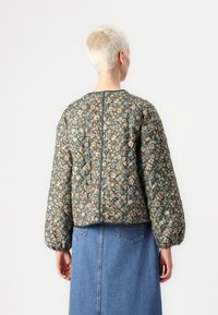 Gequilted bomberjack in bruin met een bloemenpatroon in blauw en crème. Heeft opgestoken mouwen, een ronde halslijn en een detail in de naad aan de achterkant.