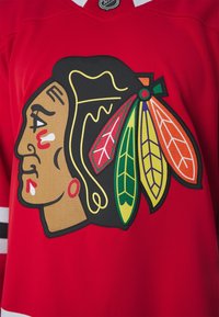 Maglia rossa con un grande e dettagliato logo tribale nero e multicolore, con piume stilizzate e un motivo di trama testurizzata.