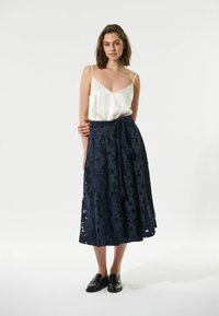 Camisola di seta bianca con spalline sottili abbinata a una gonna maxi in pizzo blu navy con motivi floreali e vita annodata. Mocassini neri.