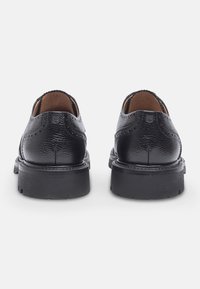Scarpe in pelle nera con finitura testurizzata, caratterizzate da dettagli brogue, un interno liscio e una suola spessa e arrotondata.