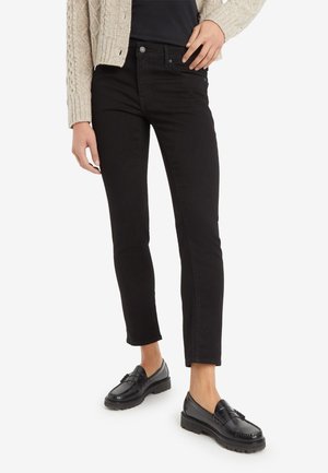 Person iført sorte slim-fit jeans, sorte loafers, sort top og beige strikket cardigan, stående mod hvid baggrund.