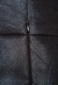 Tessuto nero texturizzato con una finitura lucida, caratterizzato da una cucitura centrale e da un piccolo dettaglio con zip. La superficie presenta un motivo leggermente in rilievo.
