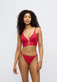 Κόκκινο σετ δαντελένιας εσωρούχων με σουτιέν βαθιάς ντεκολτέ και λεπτομέρειες με ανοίγματα, συνοδευόμενο από τανγκί, που αναδεικνύει περίπλοκα λουλουδάτα σχέδια και κυματιστά περιθώρια.