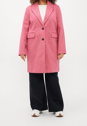 Mujer con abrigo rosa de longitud media con botones negros, pantalones negros de pierna ancha y zapatillas blancas, de pie frente a un fondo blanco.