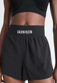 Shorts sportivi neri con una cintura ampia che presenta la scritta "CALVIN KLEIN" in bianco. Tessuto leggero, vestibilità comoda e texture liscia.