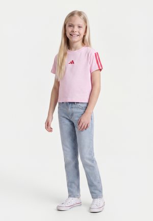 Fată zâmbitoare cu păr lung și blond, purtând un tricou roz cu mânecă scurtă, blugi albastru deschis și adidași albi, stând pe un fundal alb.