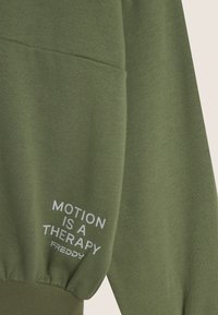 Felpa verde oliva con una trama morbida, dotata di tasca e testo stampato bianco "MOTION IS A THERAPY FREDDY" sulla manica.