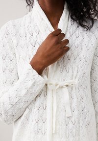 Accappatoio bianco lavorato a maglia con motivo a diamante e tessuto testurizzato. Presenta un collo a scialle e un fiocco legato in vita. Sono visibili le maniche lunghe.