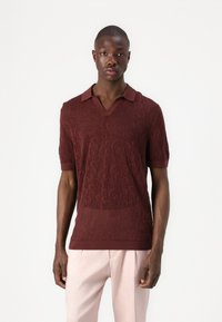 Polo shirt bordeaux à manches courtes avec un motif en dentelle texturée, col en V, et col et poignets contrastés. Porté avec un pantalon rose pâle.