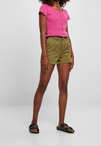 Różowy krótki top zapinany na guziki z falbanowanymi rękawkami, w zestawieniu z oliwkowozielonymi szortami z elastycznym pasem i podwiniętymi mankietami. Czarne klapki.