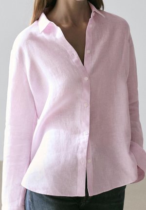 Femme portant une chemise rose clair à manches longues avec col et boutons, et un jean bleu, debout devant un fond uni.