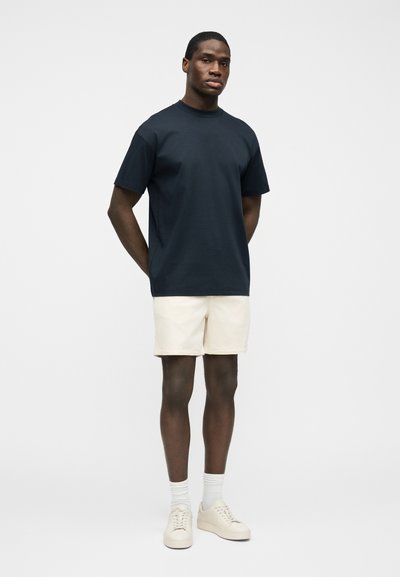 Homme debout devant un fond uni, portant un t-shirt bleu marine, un short crème, des chaussettes blanches et des baskets crème, les mains derrière le dos.