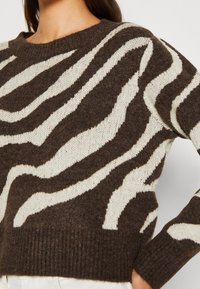 Woman wearing brown and white zebra-striped knit sweater with ribbed neckline, cuffs, and hem, paired with white pants.