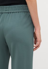 Hallivärvi rohelised joggingupüksid sujuva tekstuuriga, elastse vöökohtiga ja vabalt langeva lõikega, vaadatuna tagantpoolt käsi küljel.