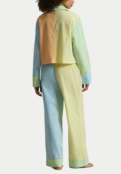 Femme portant une tenue texturée aux blocs de couleur bleu, jaune, vert et orange, debout pieds nus, de dos.