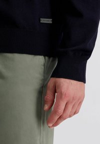 Marineblauer Strickpullover mit gerippten Bündchen und Saum, kombiniert mit hellgrünen Hosen. Besitzt ein kleines schwarzes Etikett, das die Marke angibt.