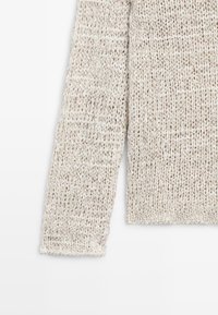 Manche longue d'un pull en grosse maille beige et blanc sur fond blanc montrant le fil texturé et le poignet côtelé.