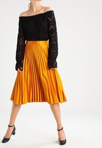 Haut noir à manches longues dénudées avec des détails en dentelle, associé à une jupe plissée de longueur genou de couleur orange vif et des escarpins noirs.