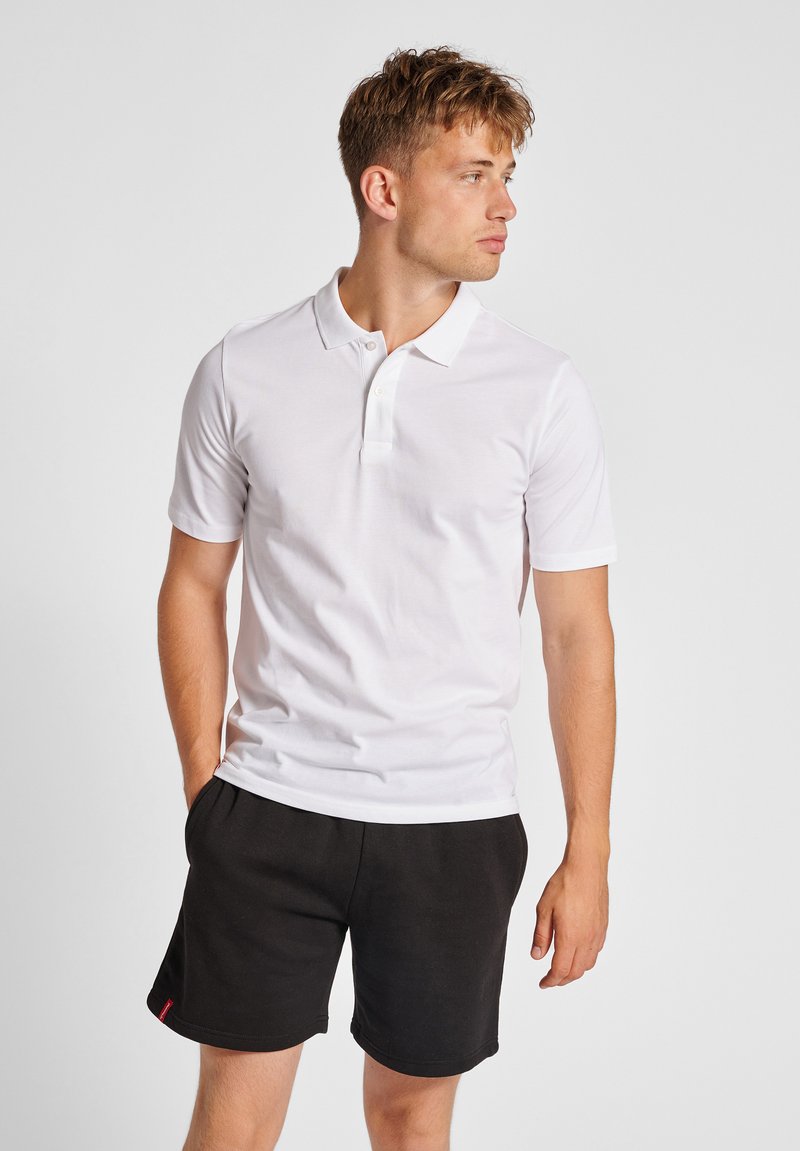 Hvid bomulds polo t-shirt med korte ærmer, flad krave og treknapslukning, parret med sorte shorts. Simpelt design, ingen mønstre.