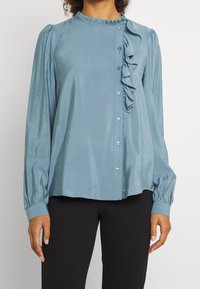 Light blue blouse with a round neckline, front button closure, and ruffle detail. Made of a smooth fabric with long, gathered sleeves.