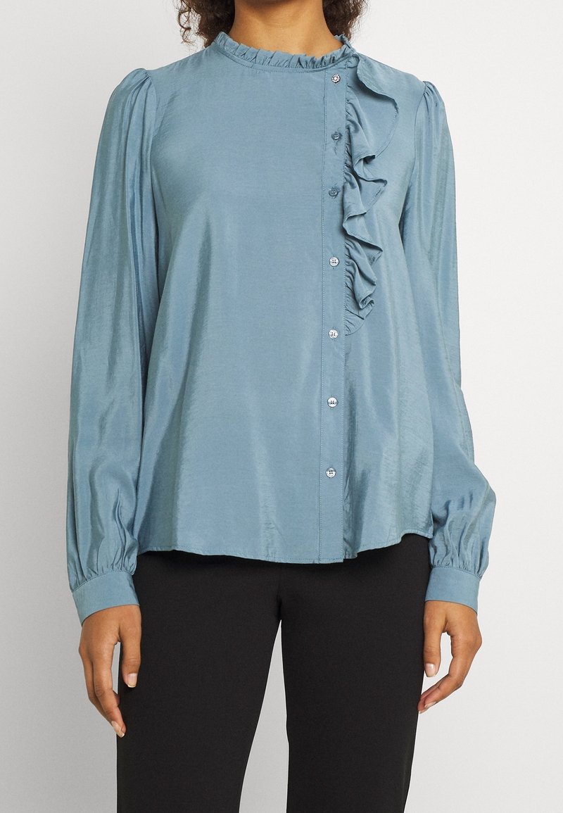 Light blue blouse with a round neckline, front button closure, and ruffle detail. Made of a smooth fabric with long, gathered sleeves.