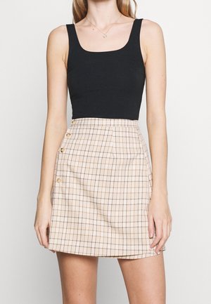 Kobieta nosząca czarny, bezrękawnikowy crop top w połączeniu z beżową i brązową kratowaną mini spódnicą na guziki.