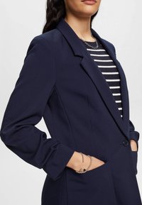 Marinblå kavaj i mjukt, texturerat tyg med en enkel knappstängning, slagkrage och fickor med klaff. Bärs över en randig topp.