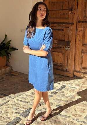 Žena v modrých šatách po kolená a sandáloch na podpätku stojaca so skríženými rukami na kamennej podlahe blízko drevených dverí.