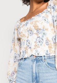 Top corto floreale con maniche lunghe a sbuffo. Presenta uno sfondo bianco con fiori blu e arancioni, corpo arricciato e orlo a balze. Abbinato a jeans azzurri chiari.