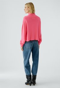 Fuchsia-Kurzpullover mit langen Ärmeln, kombiniert mit hochgeschnittenen blauen Jeans und schwarzen Stiefeletten. Glatte Textur und lockere Passform.
