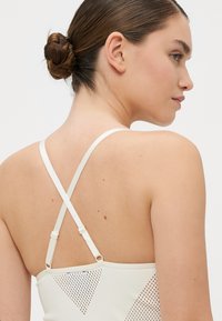Vitt sporttop med korsade band, nätinsatser och en figurnära design. Slät textur med minimala sömnadsdetaljer.