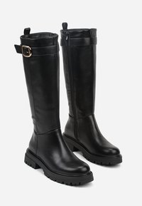 Bottes en cuir noir montantes avec une semelle épaisse, des accents en métal doré, une fermeture éclair sur le côté et un détail de boucle en haut.