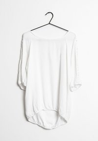 Blouse blanche à manches trois-quarts avec un décolleté arrondi, présentant un ourlet arrière incurvé et plus long, et des détails en dentelle sur les manches.