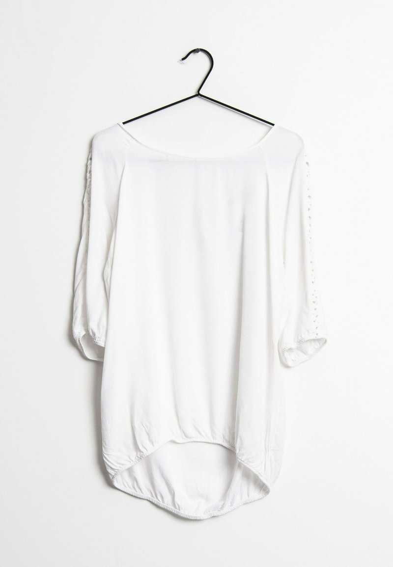 Blouse blanche à manches trois-quarts avec un décolleté arrondi, présentant un ourlet arrière incurvé et plus long, et des détails en dentelle sur les manches.