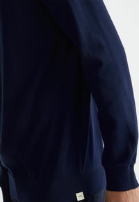 Felpa blu navy con una trama di cotone morbido, polsini e orlo a costine, con un piccolo logo in basso a sinistra.