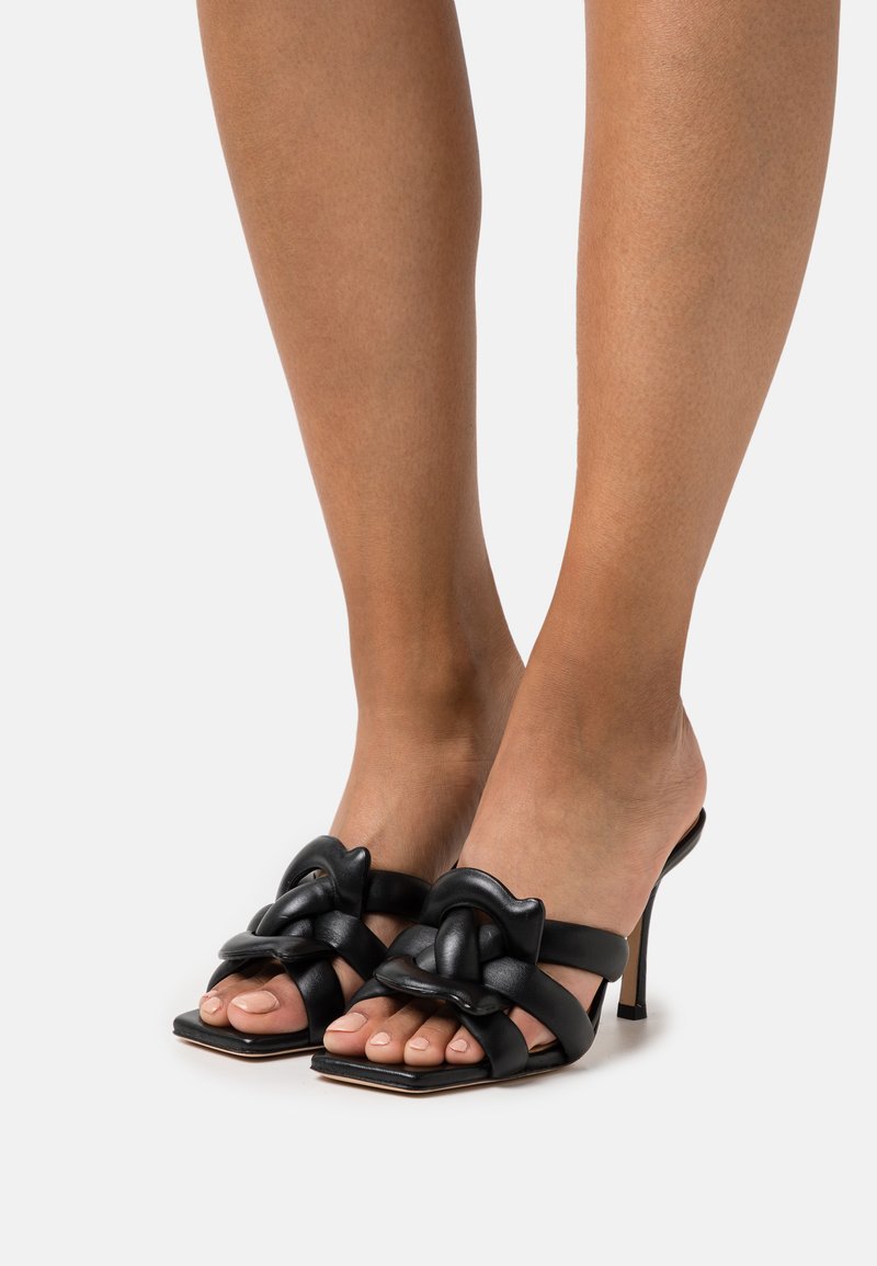 Zwarte leren hoge hakken mules met een vierkante neus, voorzien van verweven knoopaccenten en een gladde textuur.
